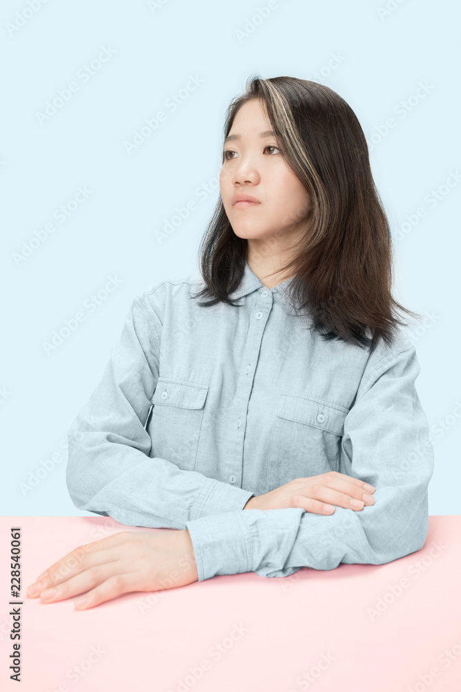 Serious business woman sitting at table, looking at left isolated on trendy blue studio background. Beautiful, young face. Female half-length portrait. Human emotions, facial expression concept. Serious business woman sitting at table, looking at left isolated on trendy blue studio background. Beautiful, young face. Female half-length portrait. Human emotions, facial expression concept.