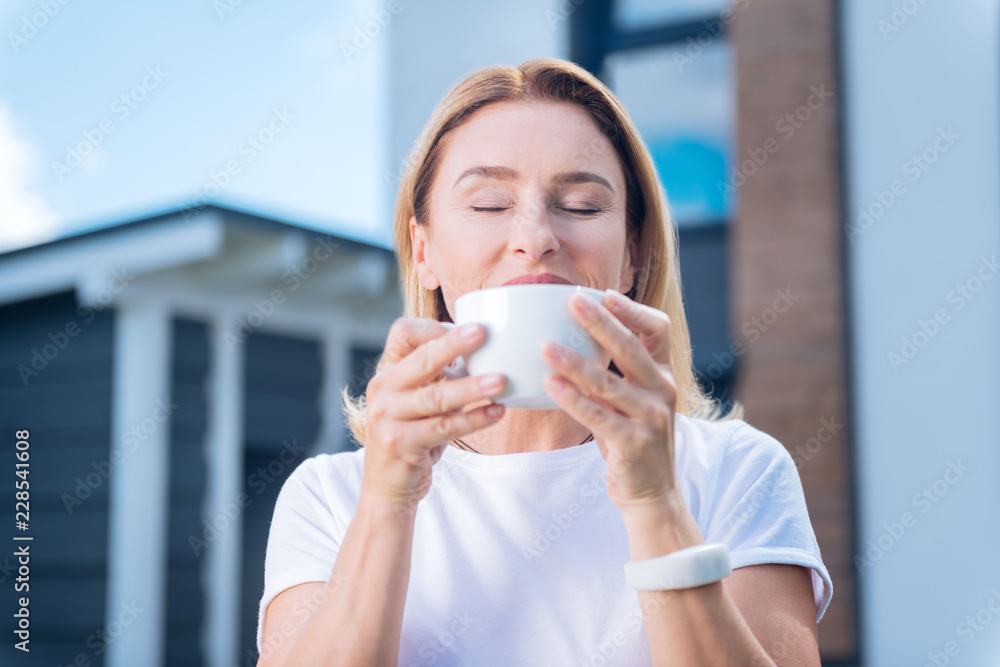 Tasting cappuccino. Beautiful mature woman tasting delicious morning cappuccino standing outside her family house