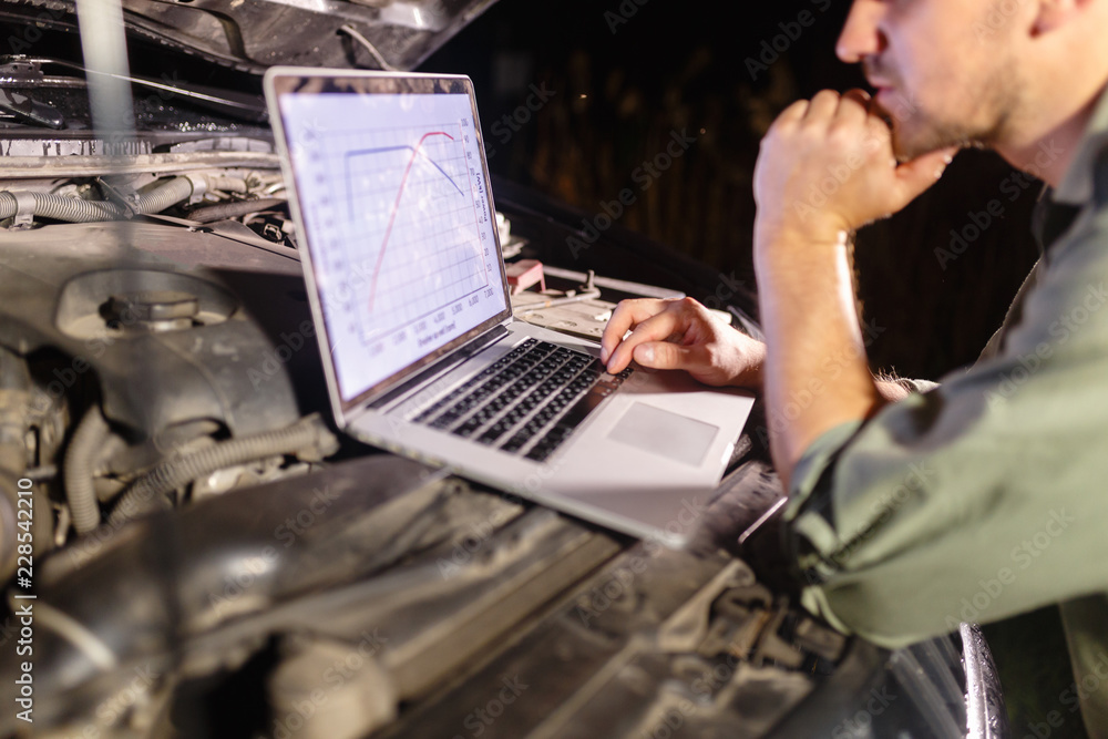 Mechanic in uniform looking intently at the laptop is connected to the motor vehicle for testing and adjustment. Mechanic carries out maintenance of the car. The removal of bugs of the engine. Mechanic in uniform looking intently at the laptop is connected to the motor vehicle for testing and adjustment. Mechanic carries out maintenance of the car. The removal of bugs of the engine.