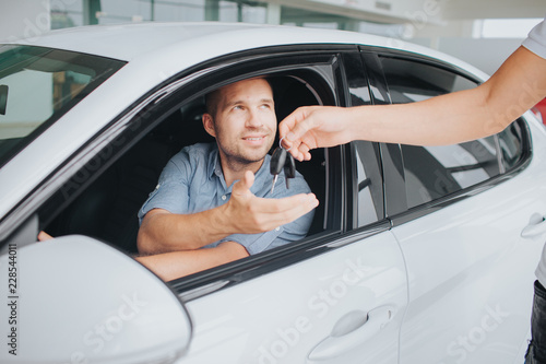 Happy owner of white car sitting inside of it and look at manager that gives key to him. Young man stretches his hand. The other one is on steering wheel. © estradaanton