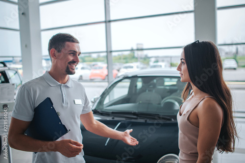 Positive and smileful man stand and point on black car. He looks at young woman. She is listening to him. Model is calm and peaceful. photo