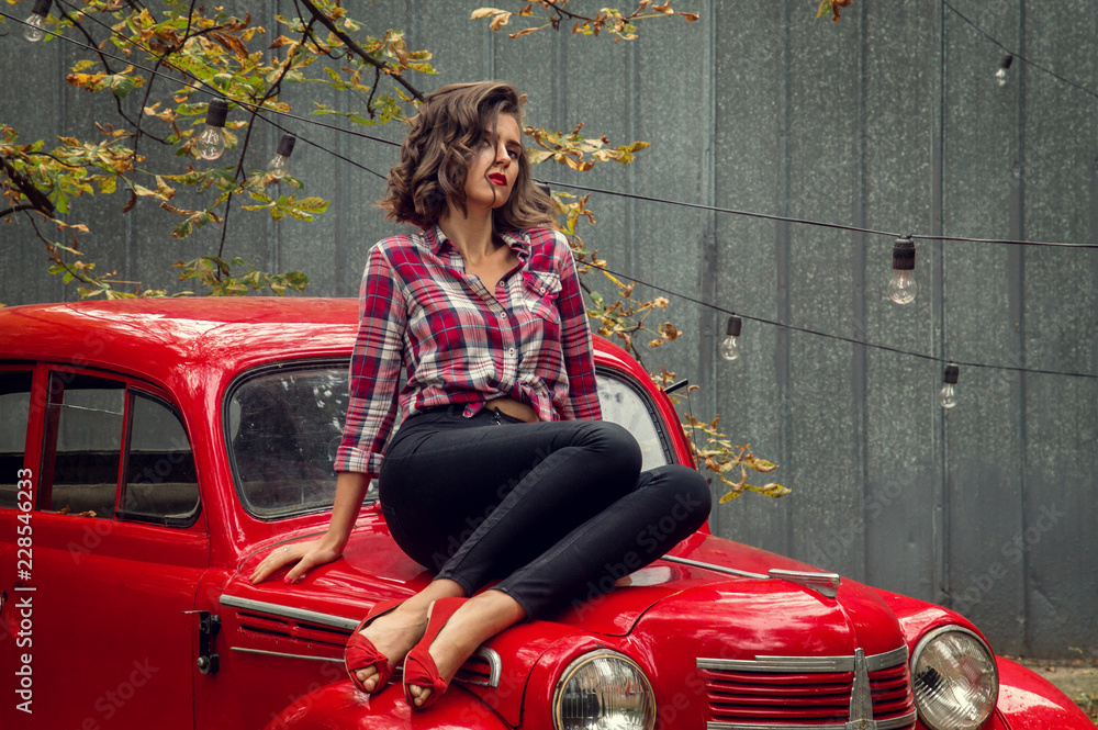 Beautiful pin-up girl in jeans and a plaid shirt posing, sitting on the  hood of
