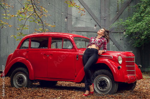 Fotografie Pin-up girl in jeans and a plaid shirt is leaning on a russian red retro car