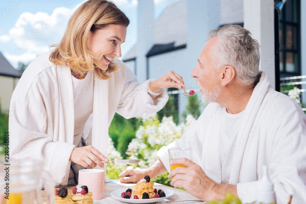 Caring wife. Beaming appealing housewife taking care of her husband during breakfast while giving him little raspberry Caring wife. Beaming appealing housewife taking care of her husband during breakfast while giving him little raspberry