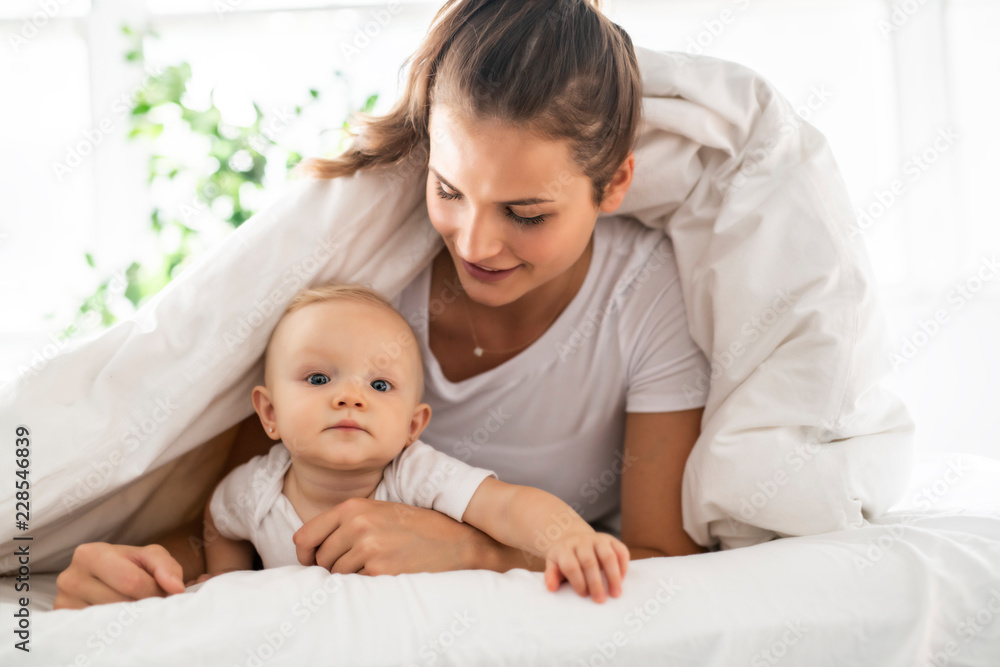 Portrait of young mother with infant baby girl liying on the bed covered with a white blanket Portrait of young mother with infant baby girl liying on the bed covered with a white blanket