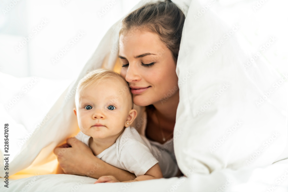 Portrait of young mother with infant baby girl liying on the bed covered with a white blanket Portrait of young mother with infant baby girl liying on the bed covered with a white blanket