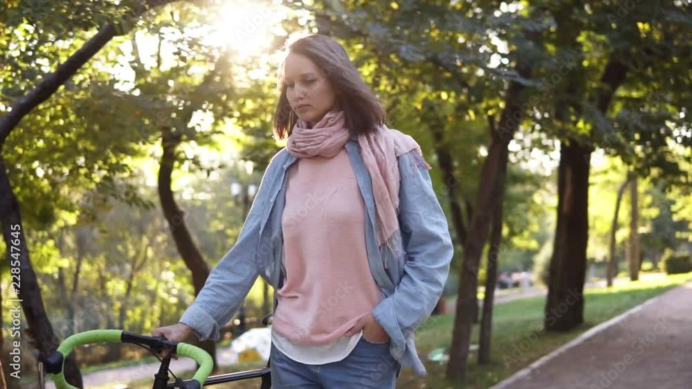 Front view of a young woman walking besides a bicycle in the morning park. The girl walking with her trekking bike, holding with a rudder. Trees and sun shines on the background. Lens flares Front view of a young woman walking besides a bicycle in the morning park. The girl walking with her trekking bike, holding with a rudder. Trees and sun shines on the background. Lens flares