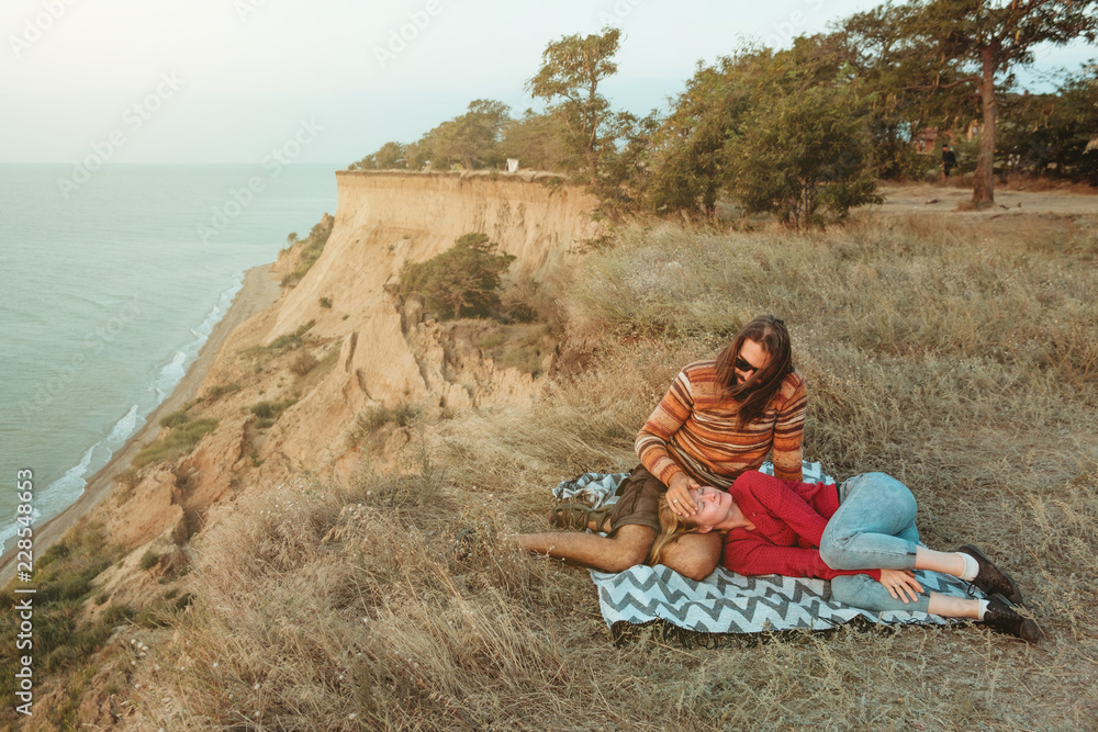 Hipster couple chilling and hugging on blanket on rocky coast with seaview