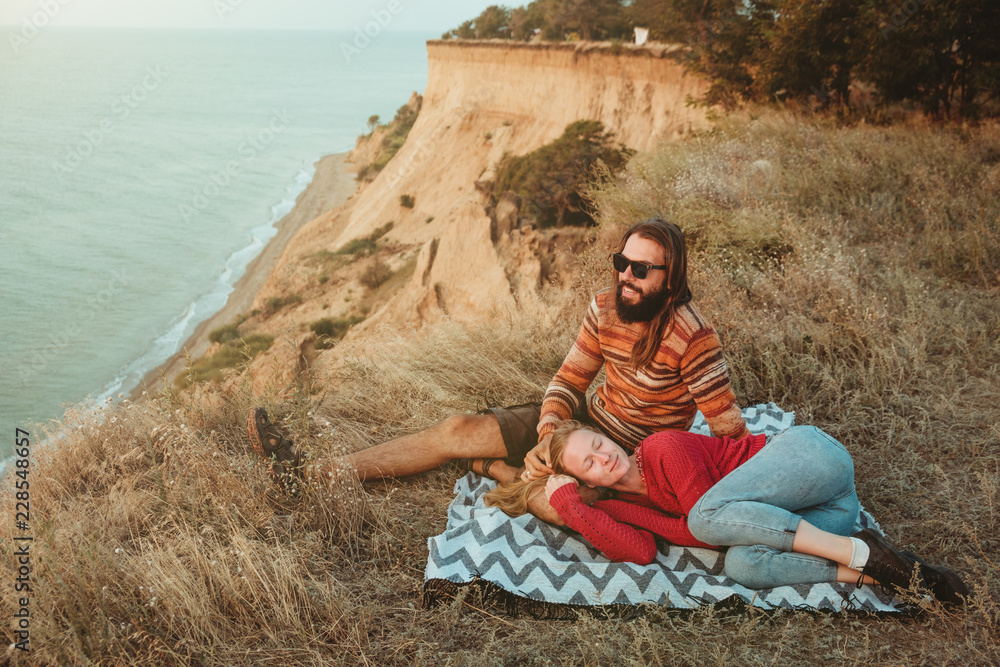 Hipster couple chilling and hugging on blanket on rocky coast with seaview