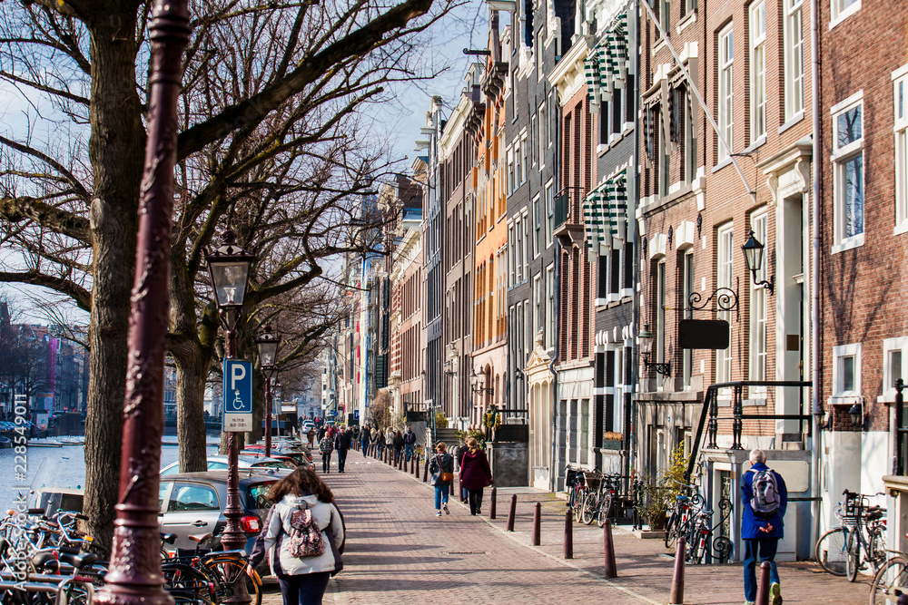 People walking and riding bicycles at the beautiful cobblestone streets next to the canals of the Old Central district of Amsterdam People walking and riding bicycles at the beautiful cobblestone streets next to the canals of the Old Central district of Amsterdam