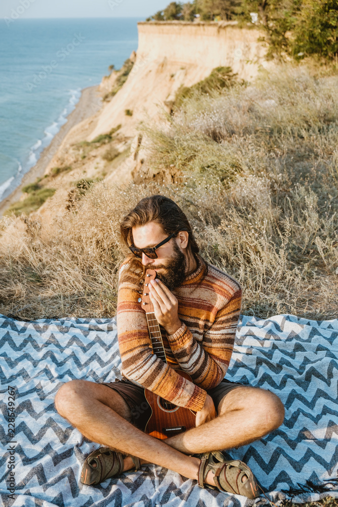 Cool guy in sweater and sunglasses playing ukulele on blanket near sea.