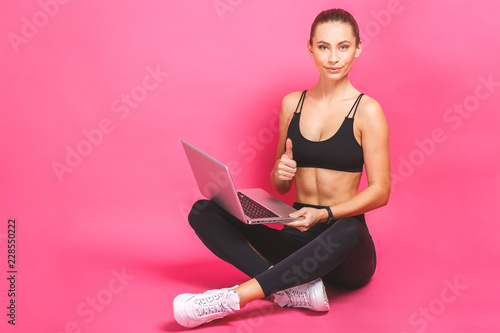 Happy beautiful fitness woman using laptop over pink background. Looking at camera, thumbs up.