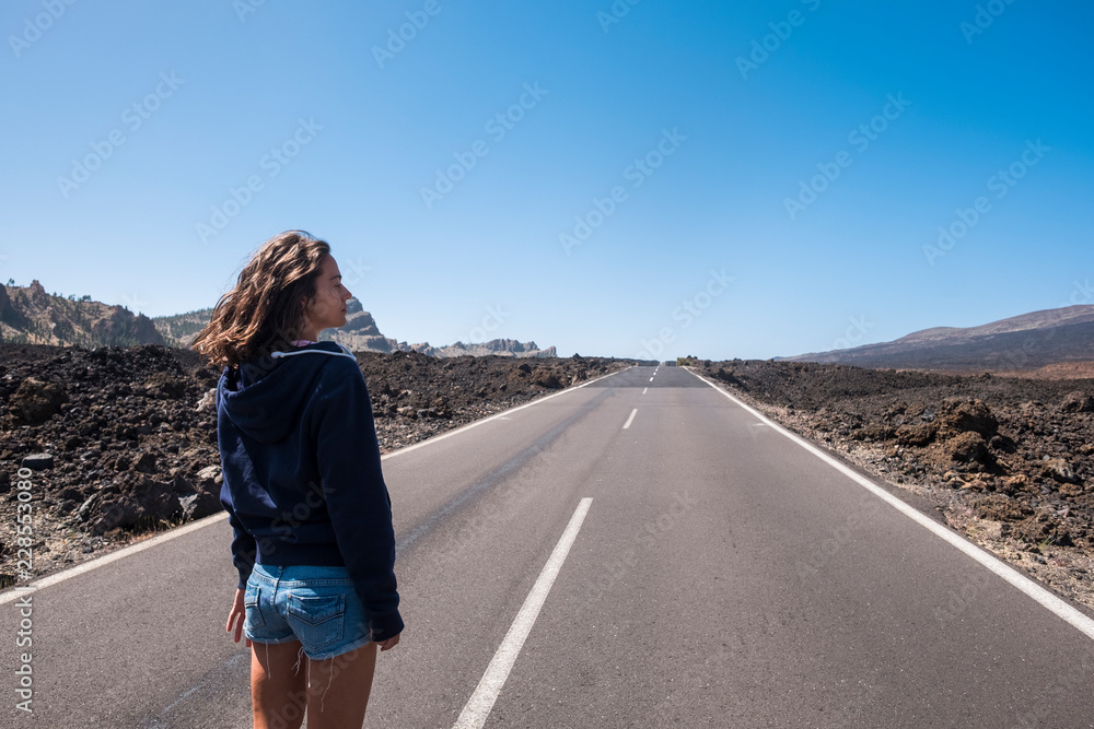 Lonely beautiful young caucasian woman standing in front of a long way straight road in the middle of the lava landscape and mountains. travel and wanderlust experience and concept for lonely traveler