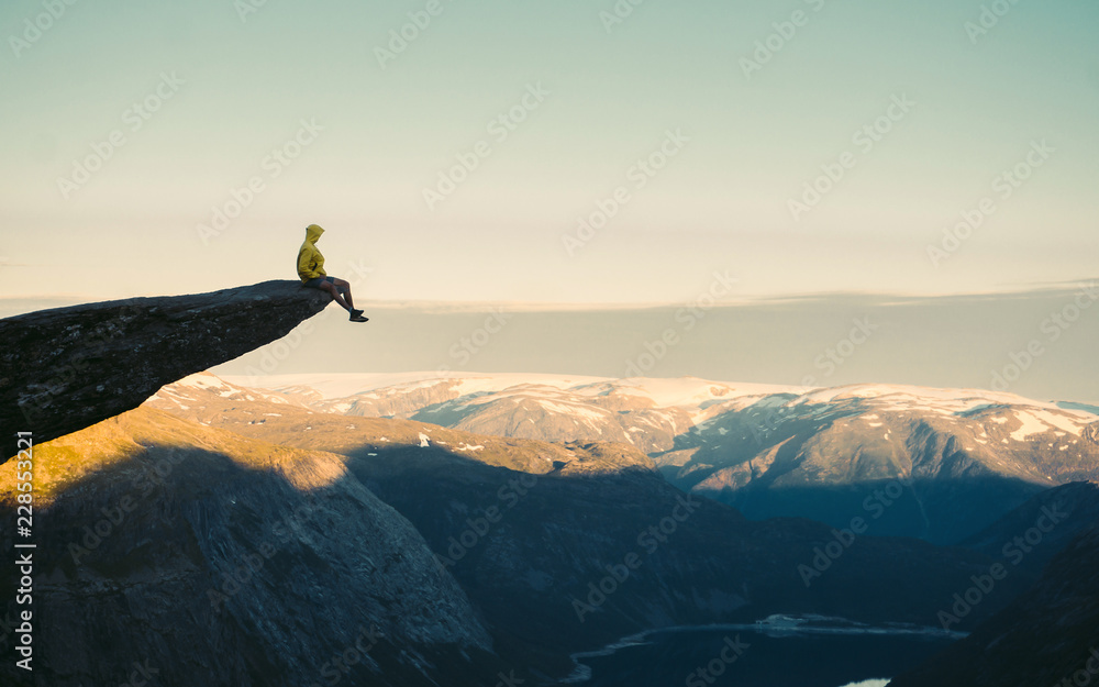 Adventurous man sitting on top of the mountain and enjoying the beautiful view during a vibrant sunset. Trolltunga rocky cliff edge in Norway mountains  - obrazy, fototapety, plakaty 