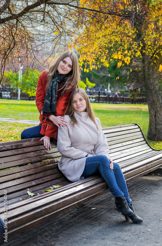 Two beautiful girlfriends sitting side by side on a bench and smile.
