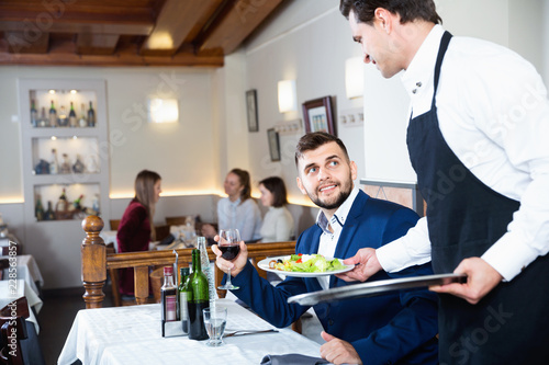 Waiter serving delicious salads to handsome young man at restaurant