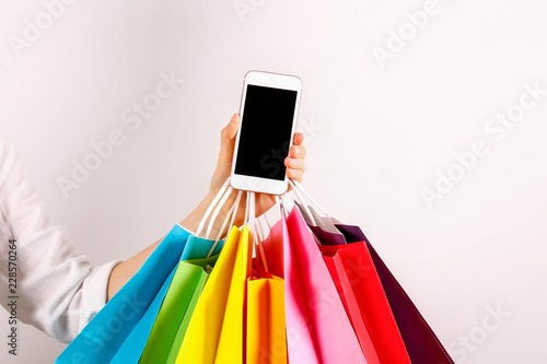 Cropped shot of female hand holding bunch of different colorful blank shopping bags and blank screen mobile phone over isolated white background. Black friday sale concept. Copy space, close up.