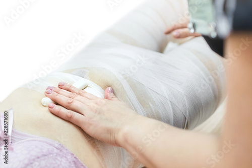 Fotografie Cosmetologist wraps a body of a woman in a white bandage