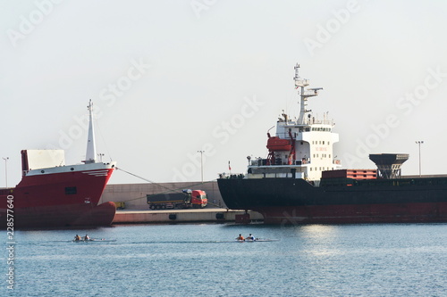 Freight truck stands between largo cargo ships in shipping harbor  oarsmen rowing in foreground sea  port Monopoli  Italy  freight digitization  transportation efficiency concept  sunny day copy space