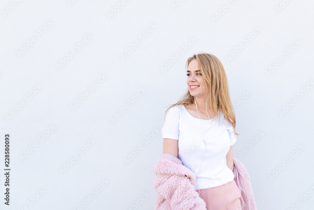 Attractive smiling girl in stylish light clothes and headphones poses on a white background and looks sideways on copyspace. Happy fashionable girl posing against the background of a white wall. Attractive smiling girl in stylish light clothes and headphones poses on a white background and looks sideways on copyspace. Happy fashionable girl posing against the background of a white wall.