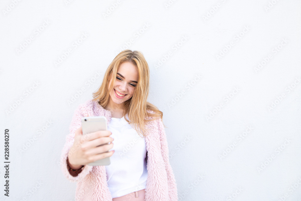 Attractive girl wearing a white T-shirt and pink coat, smiles and uses a smartphone on a white background. Cute girl with a smartphone on the background of a white wall