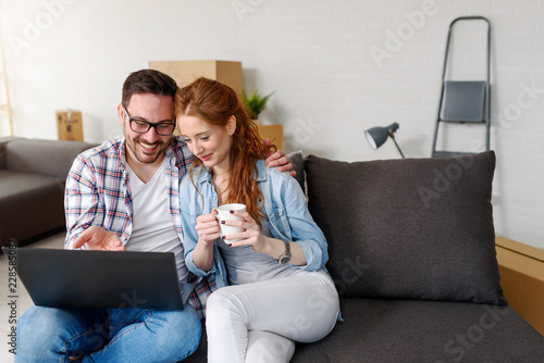Beautiful young couple  shopping on-line using a laptop and smiling while sitting among cardboard boxes in their new apartment