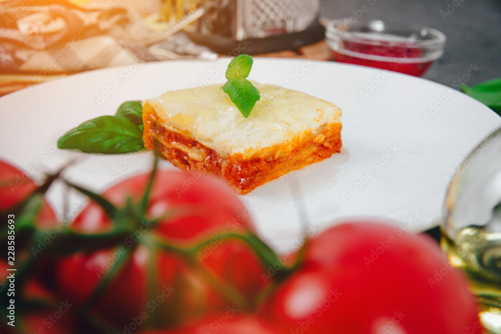 View of lasagna. A piece of lasagne on a white plate on a dark stone counter and various ingredients like basil, cheese, oil, tomatoes. The concept of preparing lasagne and pasta. View of lasagna. A piece of lasagne on a white plate on a dark stone counter and various ingredients like basil, cheese, oil, tomatoes. The concept of preparing lasagne and pasta.