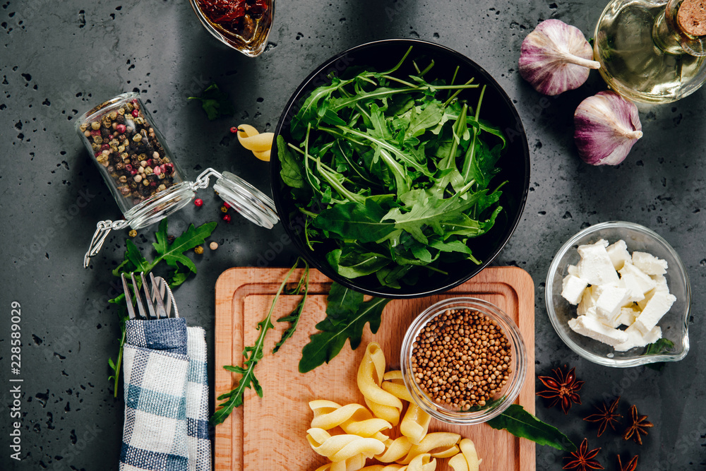 Top view of food ingredients, rucola, garlic, dried tomatoes in brine, pasta, feta cheese. The concept of preparing dishes with pasta and rucola on a stone counter top. Food photography.