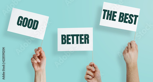 White flags with the words GOOD, BETTER, THE BEST kept in hands on a blue, pastel background. The concept of being better, developing skills, doing a better job.