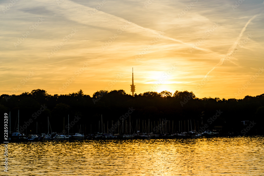 Silhouette scenery of forest tree line and waterside along Wannsee lake and television tower during sunset time and golden sky.
