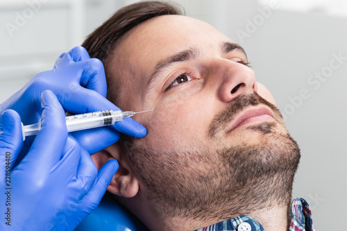 Handsome man is getting a rejuvenating facial injections. He is sitting calmly at clinic. The expert beautician is filling male wrinkles by hyaluronic acid.