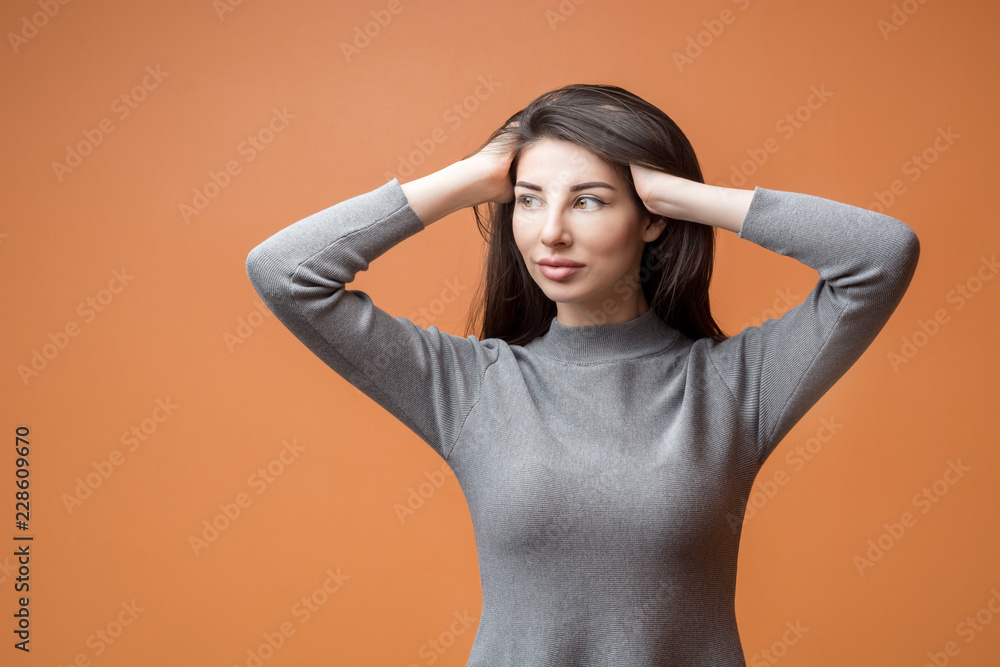 Closeup portrait of a young beautiful brunette woman with long hair holding her hands in her hair and looking to the left, thinking about something against orange background Closeup portrait of a young beautiful brunette woman with long hair holding her hands in her hair and looking to the left, thinking about something against orange background