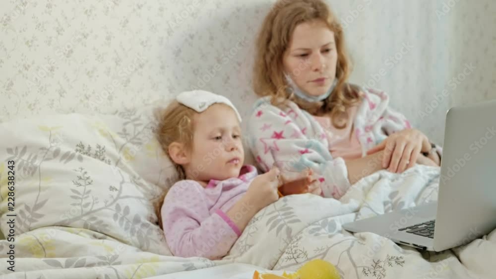 Sick girl with a temperature. Child with fever is lying in bed with her mother, eating fruit and using a laptop. Sick girl with a temperature. Child with fever is lying in bed with her mother, eating fruit and using a laptop.