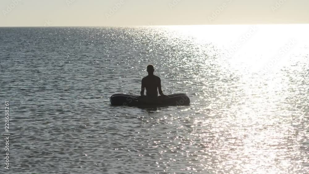 Man on an inflatable mattress in the sea is meditating, practicing yoga on swaying waves on a sunny evening. Backlit. Man on an inflatable mattress in the sea is meditating, practicing yoga on swaying waves on a sunny evening. Backlit.