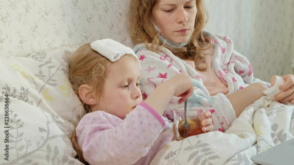 Sick girl with a temperature. Child with fever is lying in bed with her mother, eating fruit and using a laptop. Sick girl with a temperature. Child with fever is lying in bed with her mother, eating fruit and using a laptop.