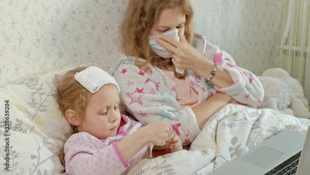 Sick girl with a temperature. Child with fever is lying in bed with her mother, eating fruit and using a laptop. Sick girl with a temperature. Child with fever is lying in bed with her mother, eating fruit and using a laptop.