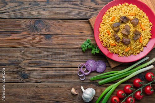 Red plate with pilaf on a brown wooden table. On the table is red pepper, green onions, garlic, cherry tomatoes, red napkin, spoon. Top view. Flat lay.