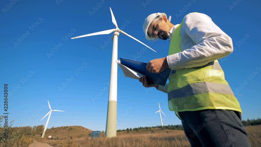 A person holds a clipboard while walking near wind turbines. Renewable electririty, green energy concept. A person holds a clipboard while walking near wind turbines. Renewable electririty, green energy concept.