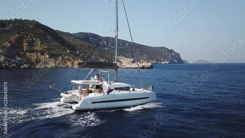 Aerial orbital view, grazing flight around a Catamaran Sailboat sailing on a deep blue sea , with rocky coast in background