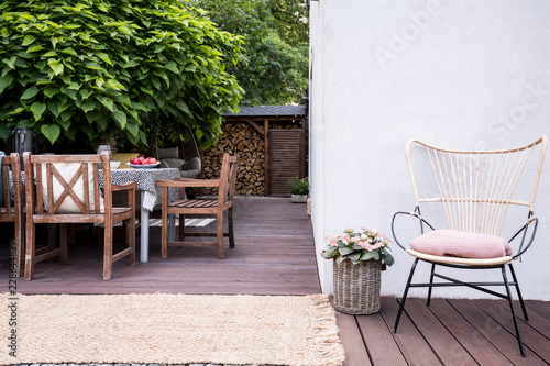 Fotografie, Obraz Pink flowers next to armchair on wooden terrace with chairs at table next to tre