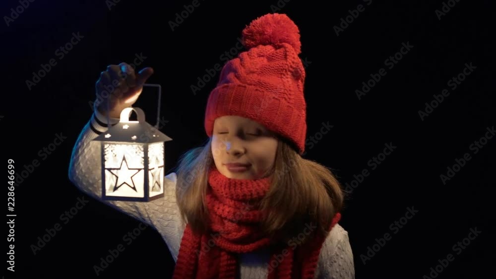 Little girl wearing knitted winter hat and scarf standing with lantern searching looking to sides with expression of surprise at the end, over black background Little girl wearing knitted winter hat and scarf standing with lantern searching looking to sides with expression of surprise at the end, over black background