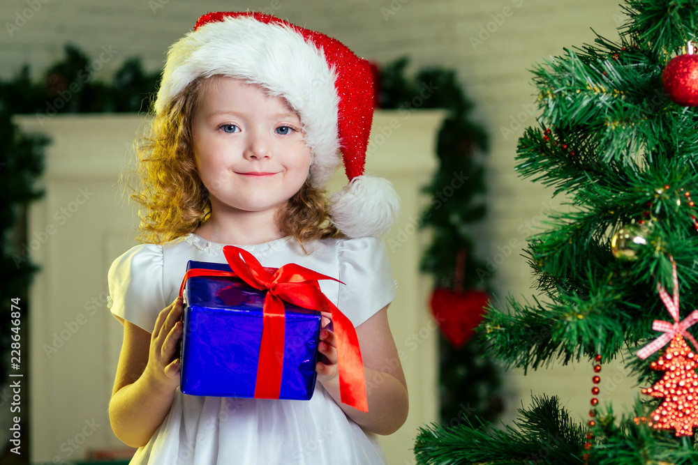 Close-up portrait of a blonde curls little girl in santa claus hat with a gift dreaming about christmas miracles standing next to the christmas tree and fireplace bright interior of the house Close-up portrait of a blonde curls little girl in santa claus hat with a gift dreaming about christmas miracles standing next to the christmas tree and fireplace bright interior of the house