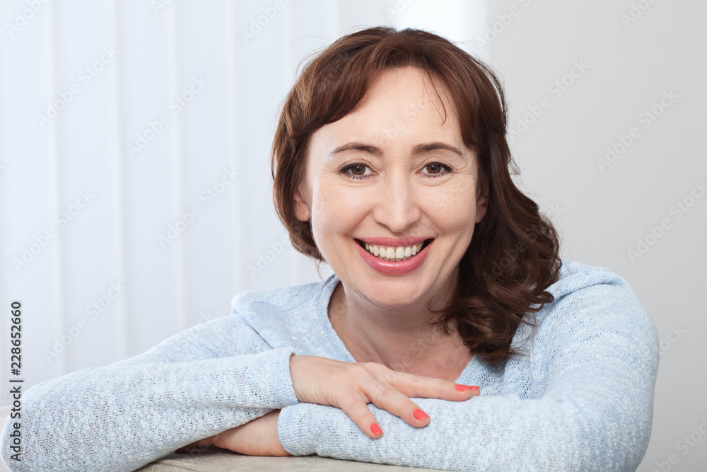 Lovely middle-aged brunette woman with a beaming smile sitting on a sofa at home looking at the camera Lovely middle-aged brunette woman with a beaming smile sitting on a sofa at home looking at the camera