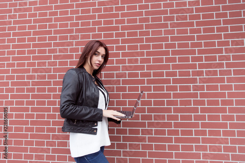 Red-haired girl in a black leather jacket works at the laptop against a red brick wall
