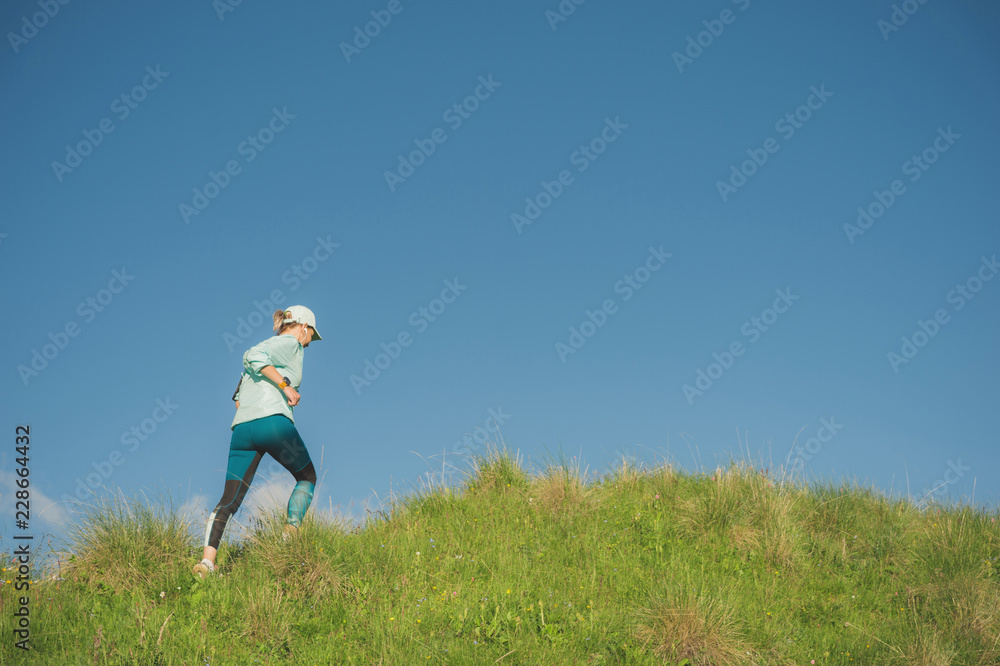 Attractive sports girl in a cap and headphones jogging through the grass in a picturesque place up the hill against the blue sky. Workout outdoors Attractive sports girl in a cap and headphones jogging through the grass in a picturesque place up the hill against the blue sky. Workout outdoors