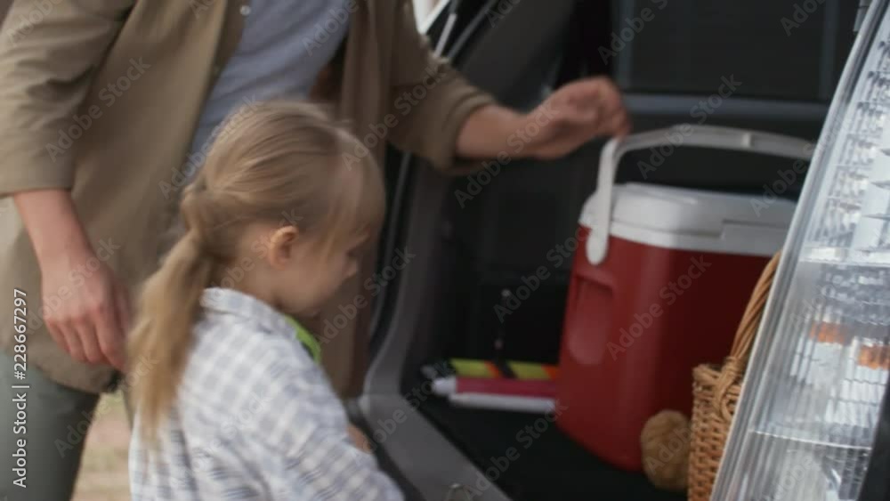 Handheld tilt down shot of bearded father and little girl packing car for picnic: they are putting cooler fridge, plush toy and vacuum flask in trunk, then walking away Handheld tilt down shot of bearded father and little girl packing car for picnic: they are putting cooler fridge, plush toy and vacuum flask in trunk, then walking away