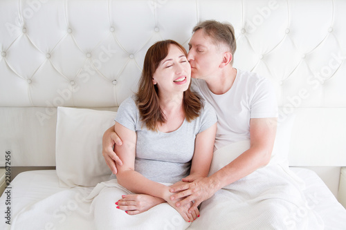 Middle age family couple in white bedroom in bed. Husband kiss wife. Love and romance. Healthy relationship. © ladyalex