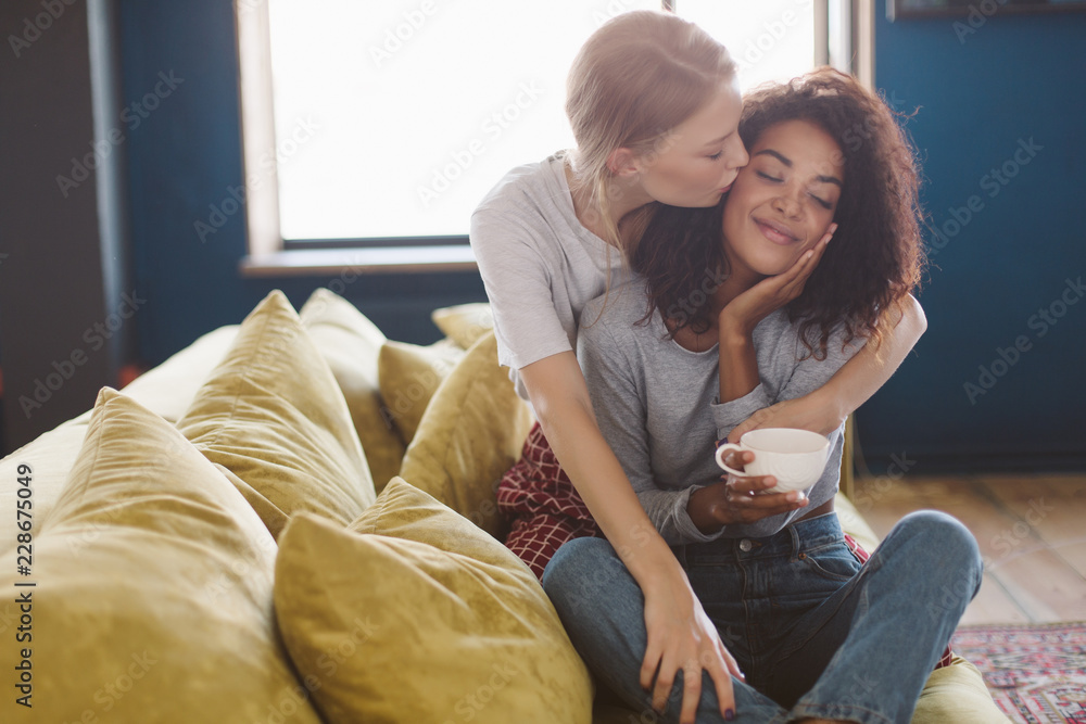 Young pretty african american girl with dark curly hair holding cup in hands dreamily closing eyes while girlfriend kissing her in cheek at cozy home Young pretty african american girl with dark curly hair holding cup in hands dreamily closing eyes while girlfriend kissing her in cheek at cozy home