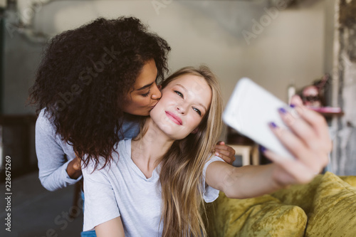 Beautiful girl with blond hair dreamily taking photos on cellphone while pretty african american girl kissing her in cheek while spending time together at home