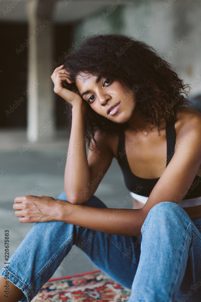 Beautiful african american girl with dark curly hair in black sporty top and jeans thoughtfully looking in camera while sitting on vintage carpet at home Beautiful african american girl with dark curly hair in black sporty top and jeans thoughtfully looking in camera while sitting on vintage carpet at home